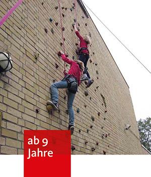 2 Kinder klettern gesichert mit Kletterseilen an einer Hauswand, an der Klettergriffe geschraubt sind