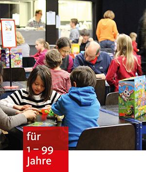 mehrere Personen, kleine und große, sitzen an Tischen und spielen